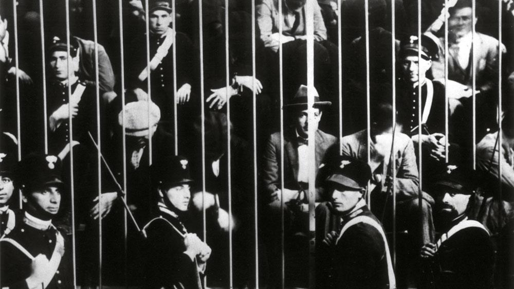 Bild: Historische Schwarz-Weiß-Aufnahme: Eine Gruppe von Männern steht dicht gedrängt hinter einem hohen Metallgitter, davor und dazwischen befinden sich mehrere uniformierte Sicherheitskräfte.