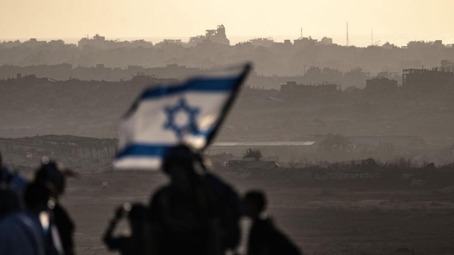 Bild: Eine unscharfe israelische Flagge weht neben Soldaten. Im Hintergrund das zerstörte Gaza. Gegen das Abendlicht fotografiert.
