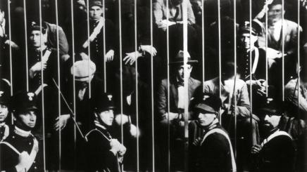 Bild: Historische Schwarz-Weiß-Aufnahme: Eine Gruppe von Männern steht dicht gedrängt hinter einem hohen Metallgitter, davor und dazwischen befinden sich mehrere uniformierte Sicherheitskräfte.