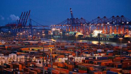 Bild: Containerhafen bei Dämmerung mit zahlreichen gestapelten Frachtcontainern, großen Hafenkränen und Containerschiffen.